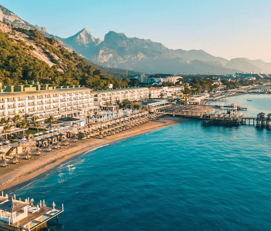 Coastal view of Corendon hotel resort with beach loungers, sandy shore, blue sea, and mountain backdrop.