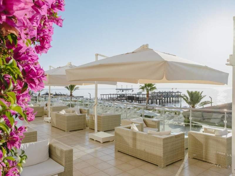 Corendon Gallery beachfront lounge with wicker seating, white umbrellas, pink bougainvillaeas, ocean view, and a wooden pier.
