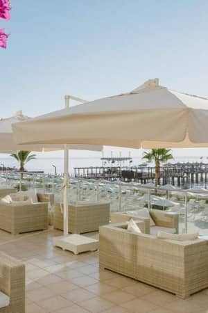 Corendon Gallery beachfront lounge with wicker seating, white umbrellas, pink bougainvillaeas, ocean view, and a wooden pier.