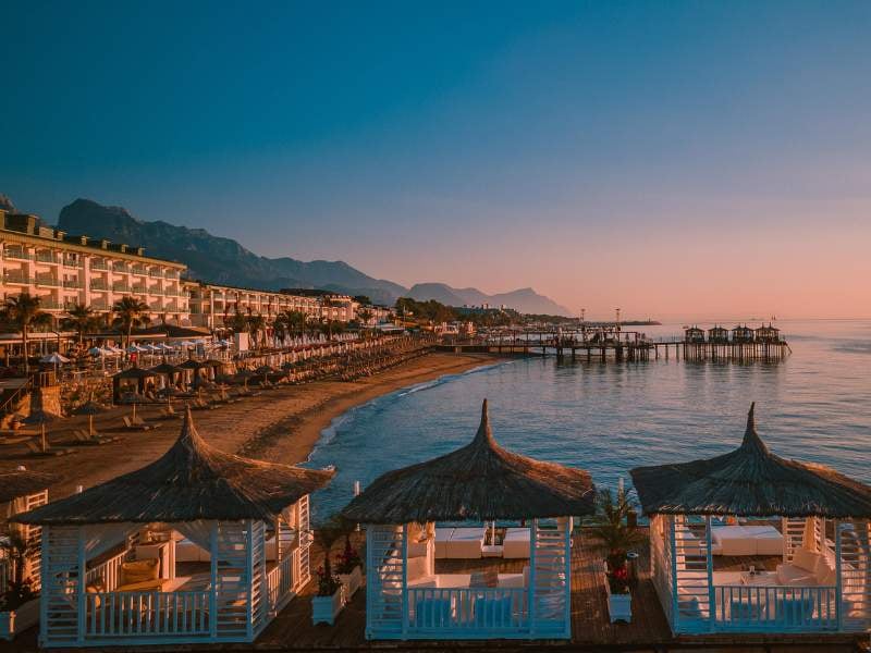 Beachfront view at sunset with luxurious straw-roofed cabanas, distant mountains, calm sea, sunbeds, and a grand hotel.