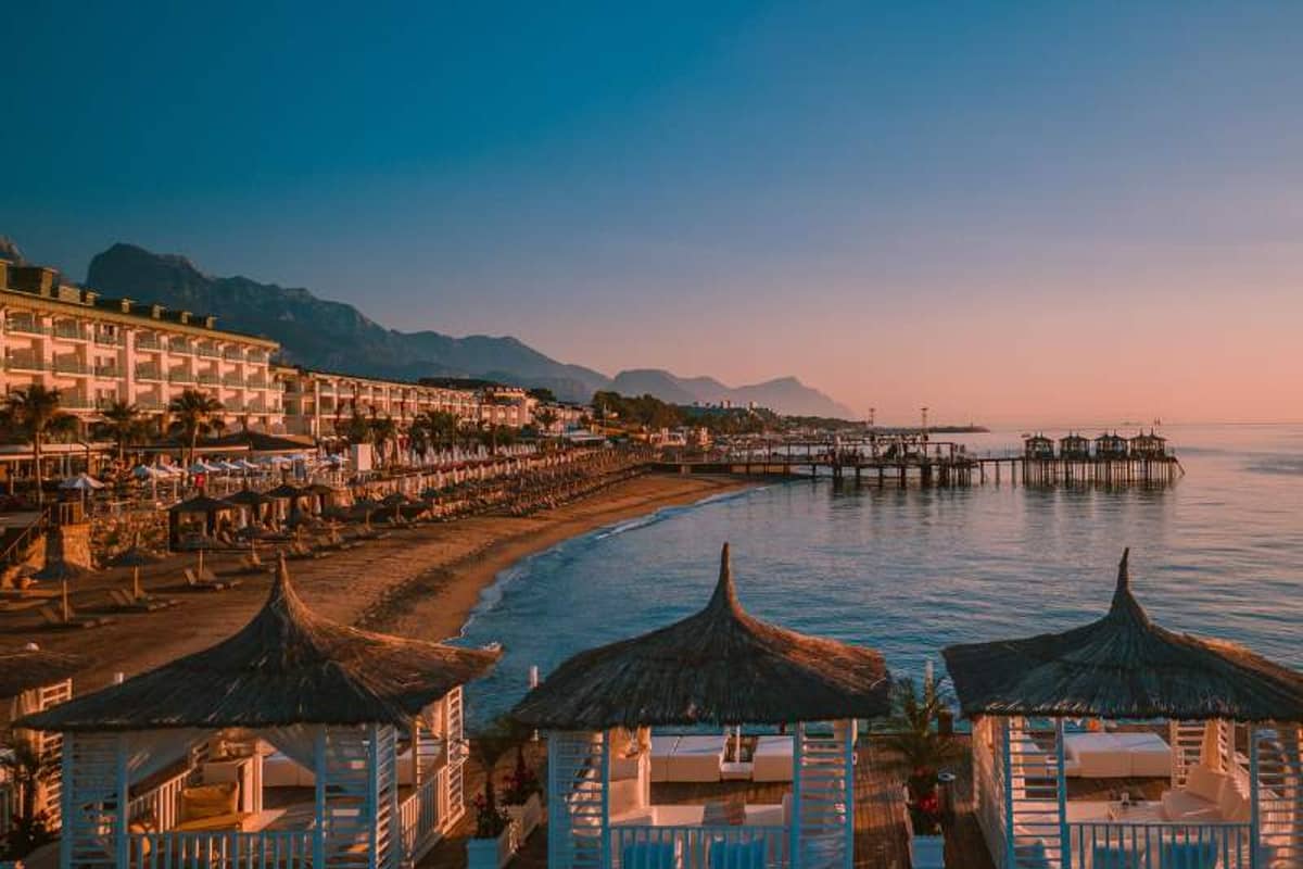 Beachfront view at sunset with luxurious straw-roofed cabanas, distant mountains, calm sea, sunbeds, and a grand hotel.