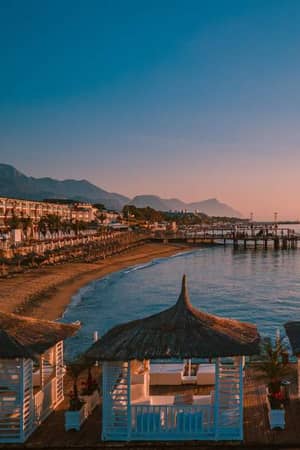 Beachfront view at sunset with luxurious straw-roofed cabanas, distant mountains, calm sea, sunbeds, and a grand hotel.