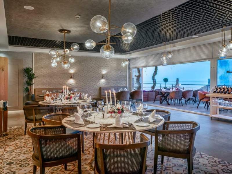 Elegant dining room at Corendon Gallery hotel with round tables, white tablecloths, and modern hanging lights, overlooking a scenic view through large windows.