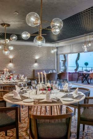 Elegant dining room at Corendon Gallery hotel with round tables, white tablecloths, and modern hanging lights, overlooking a scenic view through large windows.