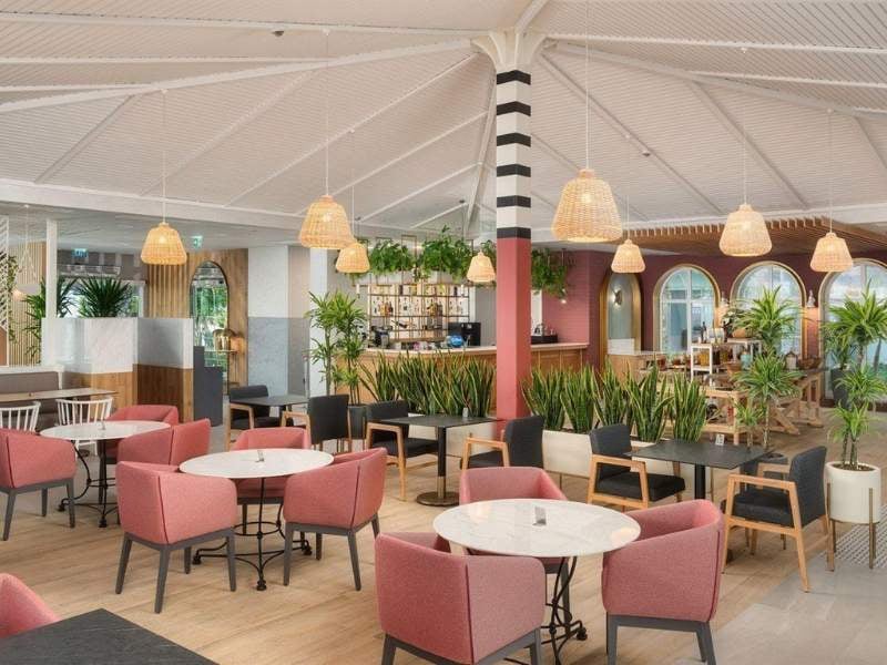 Indoor dining area at Corendon Hotel with pink chairs, round tables, potted plants, and wicker pendant lights under a vaulted ceiling.