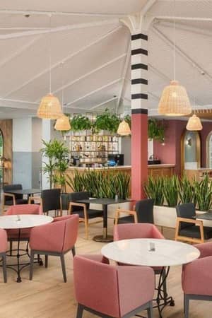 Indoor dining area at Corendon Hotel with pink chairs, round tables, potted plants, and wicker pendant lights under a vaulted ceiling.