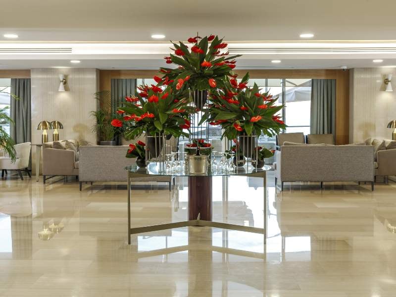 Luxurious hotel lobby with red flowers on a glass table, grey sofas, and large windows.