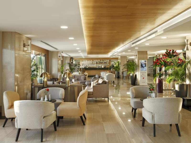 Spacious lounge with modern beige armchairs, wooden ceiling, and indoor plants at Corendon Gallery.