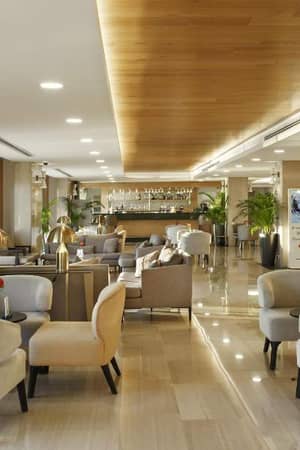 Spacious lounge with modern beige armchairs, wooden ceiling, and indoor plants at Corendon Gallery.