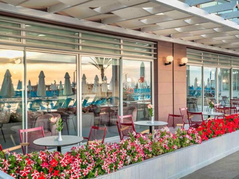 Outdoor dining area at Corendon hotel with red chairs, flower planters, and views of sun umbrellas and palm trees at sunset.