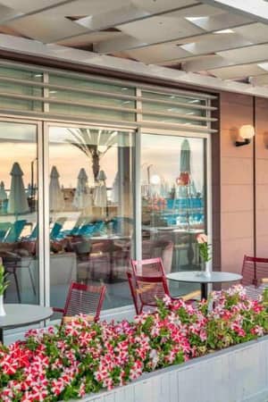 Outdoor dining area at Corendon hotel with red chairs, flower planters, and views of sun umbrellas and palm trees at sunset.
