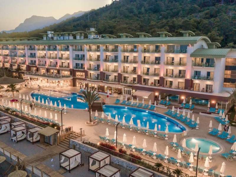 Aerial view of Corendon resort with a large swimming pool, sun loungers, umbrellas, and a hotel building set against green hills.