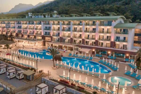 Aerial view of Corendon resort with a large swimming pool, sun loungers, umbrellas, and a hotel building set against green hills.