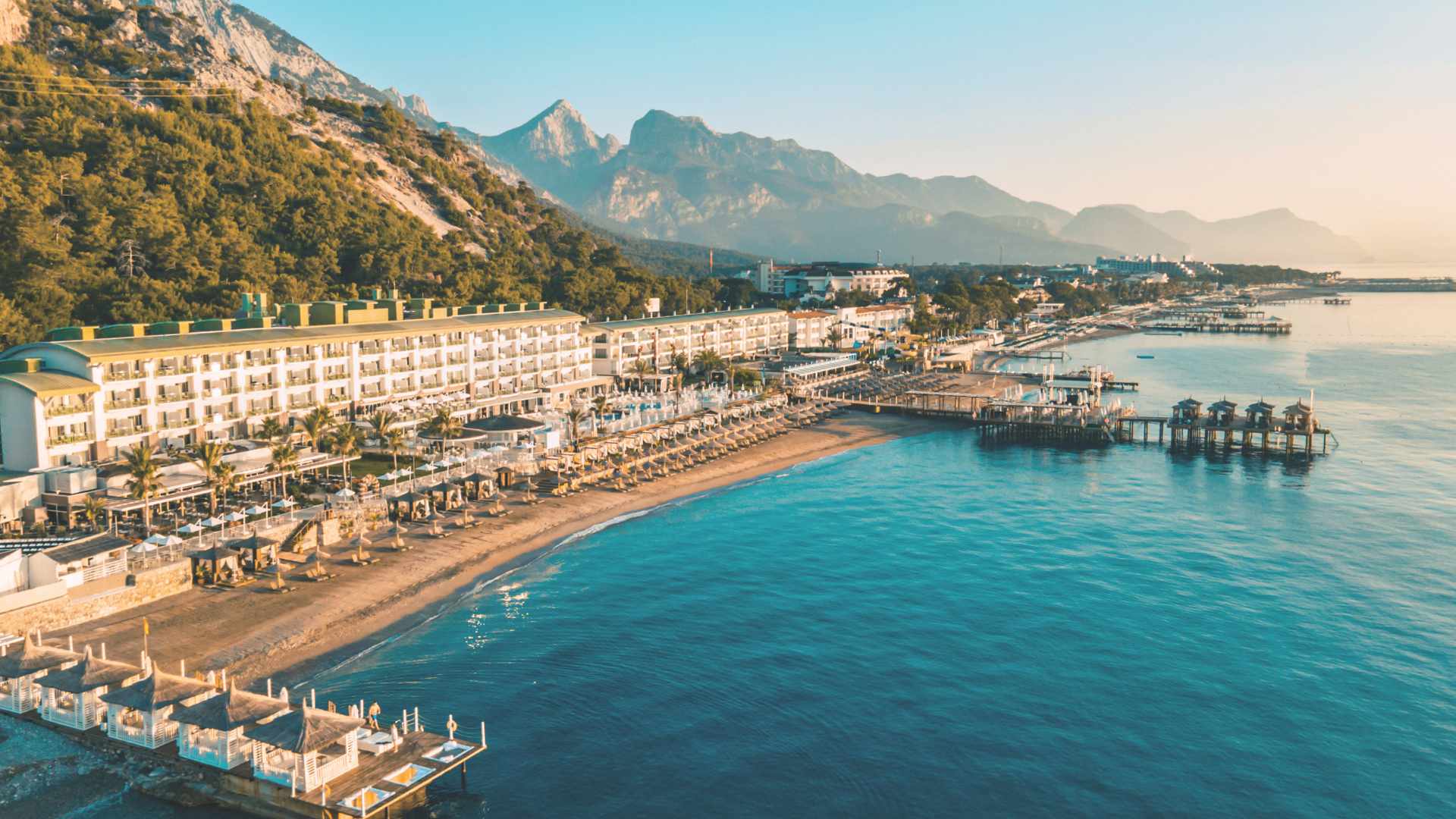 Aerial shot of a beachfront resort with a row of sunloungers and umbrellas, mountains in the background and a calm turquoise sea.