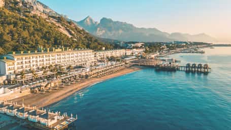 Aerial view of a beachfront resort with a large hotel, sunbeds, piers, and mountains in the background during sunset.