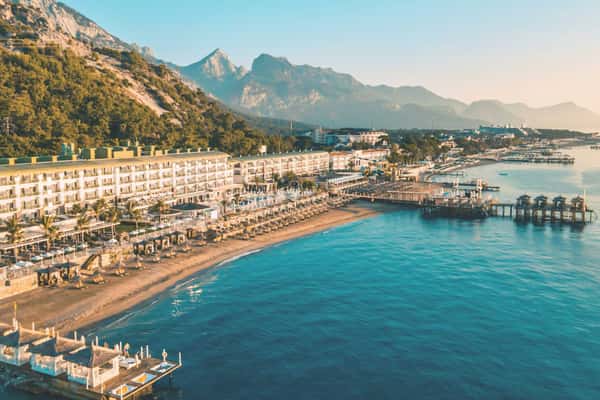 Aerial view of a beachfront resort with a large hotel, sunbeds, piers, and mountains in the background during sunset.
