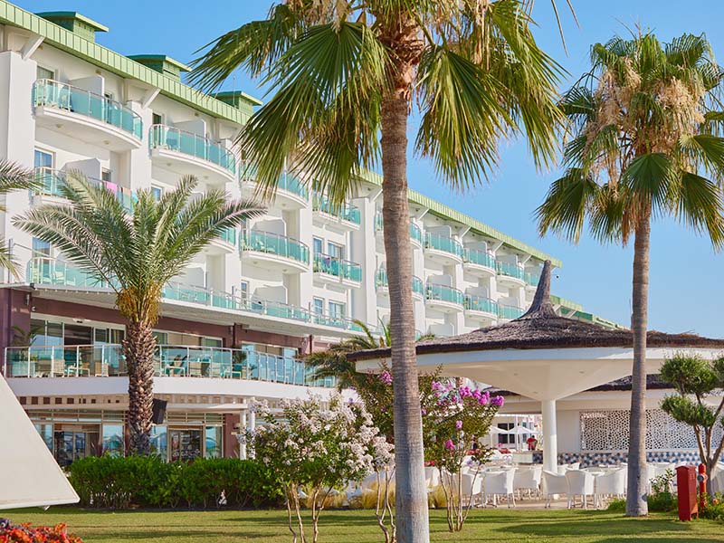 Front view of Corendon Playa Kemer Resort, showcasing its modern architecture, balconies, and surrounding palm trees and flowers.