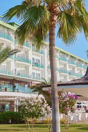 Front view of Corendon Playa Kemer Resort, showcasing its modern architecture, balconies, and surrounding palm trees and flowers.