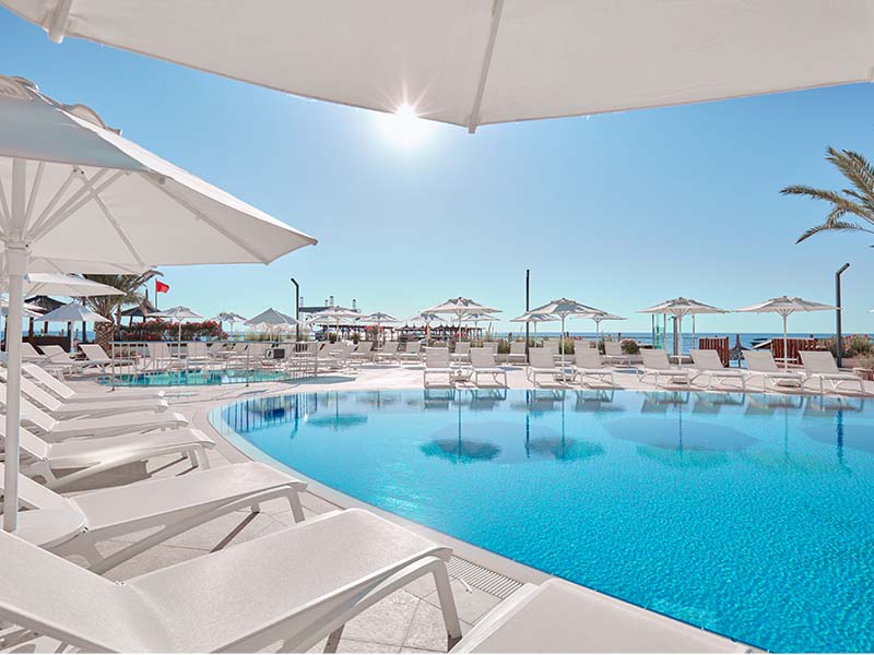 Relaxing poolside scene at Corendon Playa Kemer, featuring sun loungers, umbrellas, and a view of the sea under a clear blue sky.
