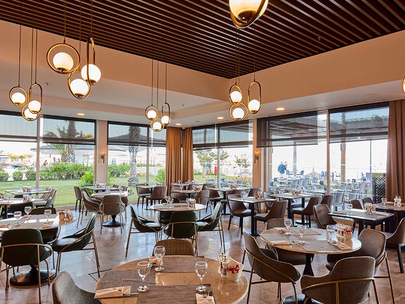 Modern dining area at Corendon Playa Kemer with round tables, pendant lights, and large windows overlooking gardens and terrace.