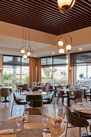 Modern dining area at Corendon Playa Kemer with round tables, pendant lights, and large windows overlooking gardens and terrace.