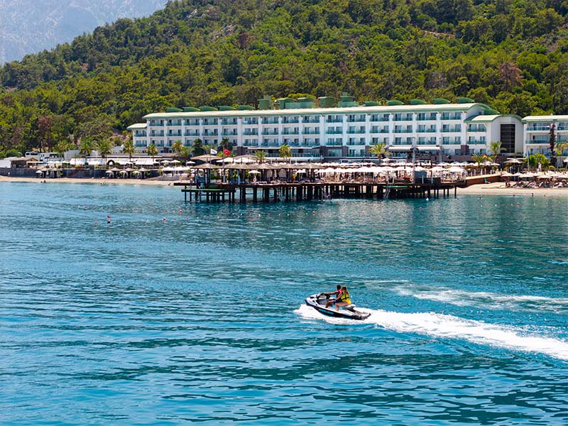 View of Corendon Playa Kemer with a lush hillside and a jet ski on turquoise waters in front of the beachfront resort.