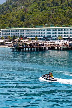 View of Corendon Playa Kemer with a lush hillside and a jet ski on turquoise waters in front of the beachfront resort.