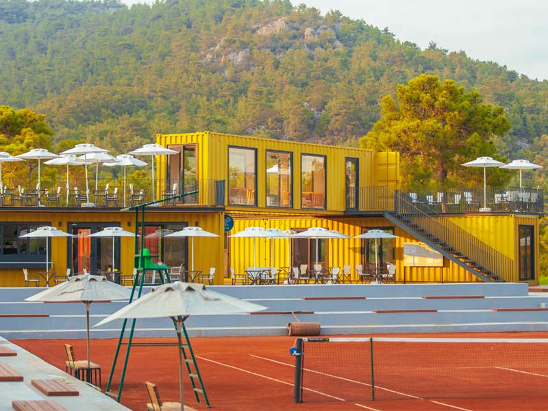 Vibrant yellow shipping container building at Corendon Playa Kemer with glass windows, outdoor seating, and lush hills in the background.