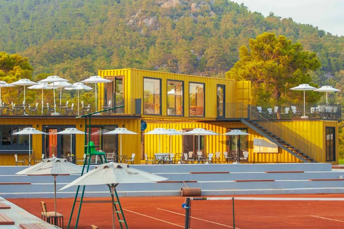 Vibrant yellow shipping container building at Corendon Playa Kemer with glass windows, outdoor seating, and lush hills in the background.
