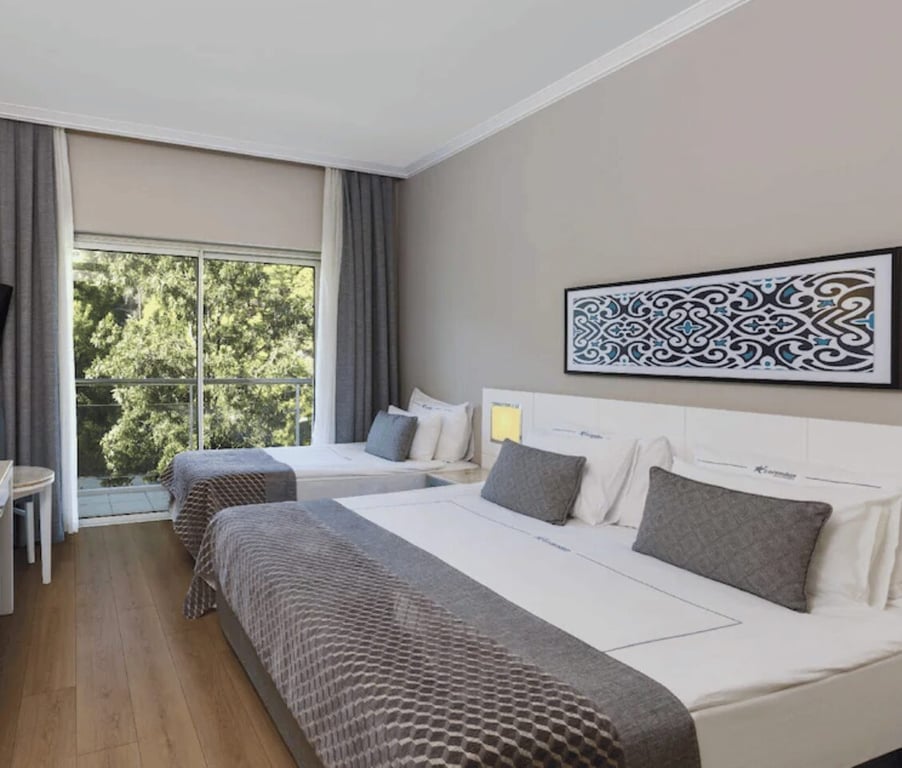 Hotel room with two double beds, grey and white bedding, a large window revealing greenery, and a decorative artwork above the beds.