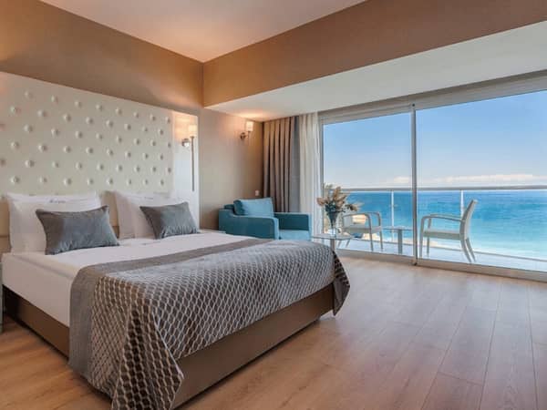 Luxurious hotel room with sea view at Corendon, featuring a double bed, beige headboard, armchair, balcony, and ocean scenery.