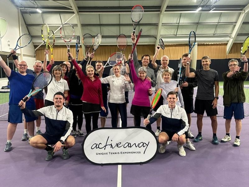 Group of tennis players at DL Bristol Gallery holding up racquets, participating in an Active Away event on an indoor court.