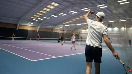 Indoor tennis court with players in a doubles match; a person in a white t-shirt serves the ball.