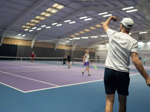 Indoor tennis court with players in a doubles match; a person in a white t-shirt serves the ball.