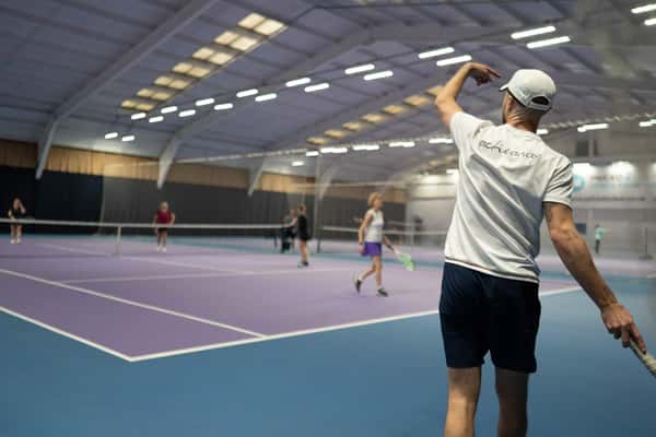 Indoor tennis court with players in a doubles match; a person in a white t-shirt serves the ball.
