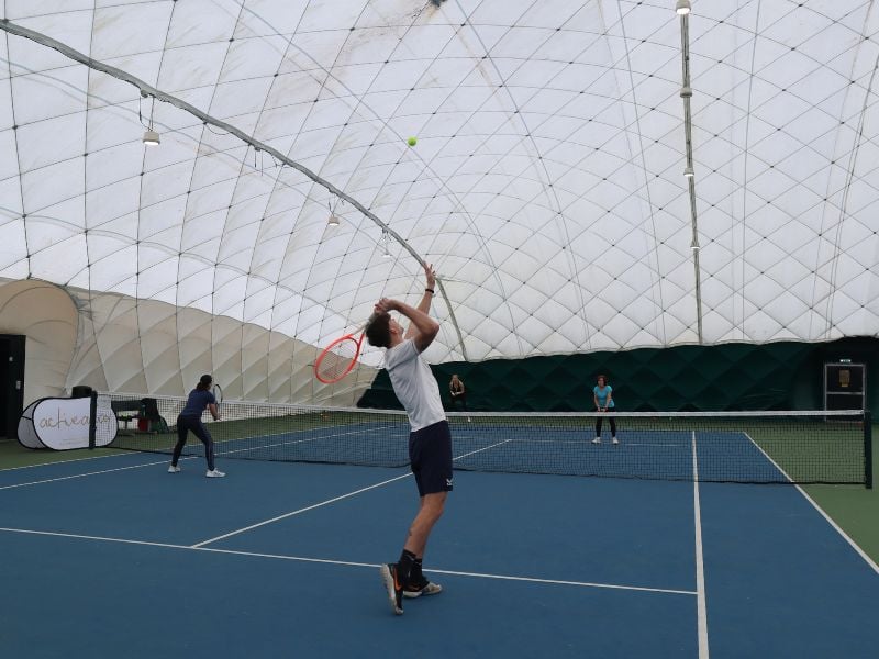 Indoor tennis coaching session in Beaconsfield, with a coach demonstrating a serve to players on the court.