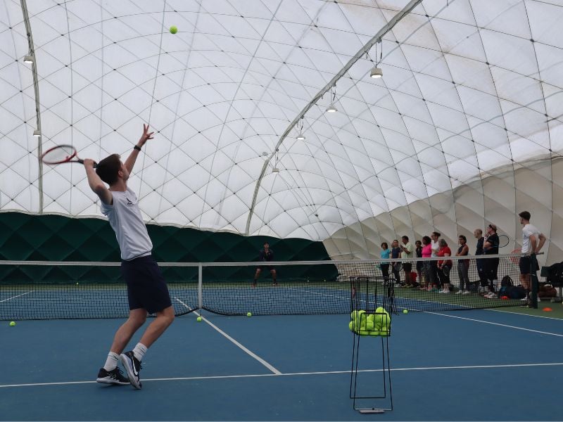 Tennis coach at DL Beaconsfield demonstrating a serve on an indoor court while a group of players observe.