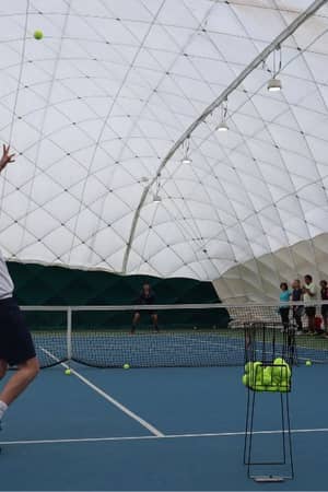 Tennis coach at DL Beaconsfield demonstrating a serve on an indoor court while a group of players observe.