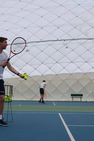Tennis coaches on court under a dome, feeding tennis balls, with Active Away logo visible, indoors at Beaconsfield.