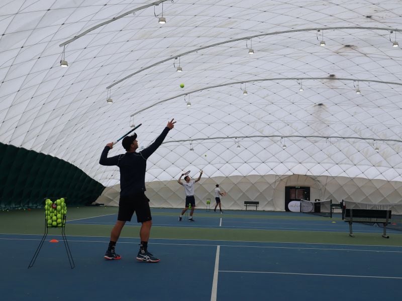 Tennis coaches serving on indoor bubble courts at David Lloyd Beaconsfield, with a basket of tennis balls and court equipment.