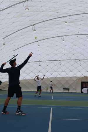 Tennis coaches serving on indoor bubble courts at David Lloyd Beaconsfield, with a basket of tennis balls and court equipment.