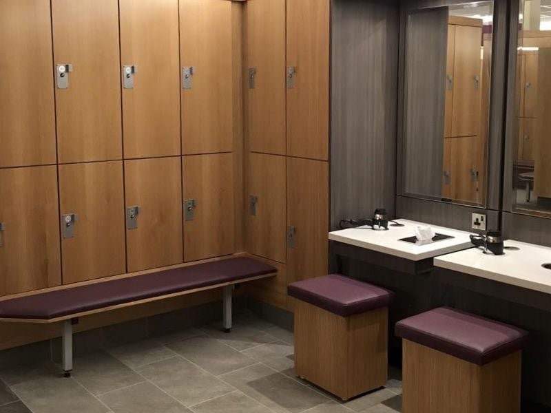 Modern changing room at DL-Beaconsfield with wooden lockers, upholstered seating, and a vanity area with mirrors and basins.