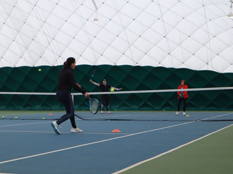 Three people on an indoor tennis court at David Lloyd Beaconsfield performing tennis drills; one player about to hit a ball with forehand stroke, two others standing near the net.