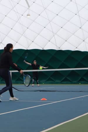 Three people on an indoor tennis court at David Lloyd Beaconsfield performing tennis drills; one player about to hit a ball with forehand stroke, two others standing near the net.