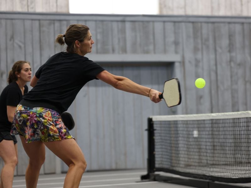 Two female pickleball players in an indoor court, with the foreground player making a forehand shot towards the net.