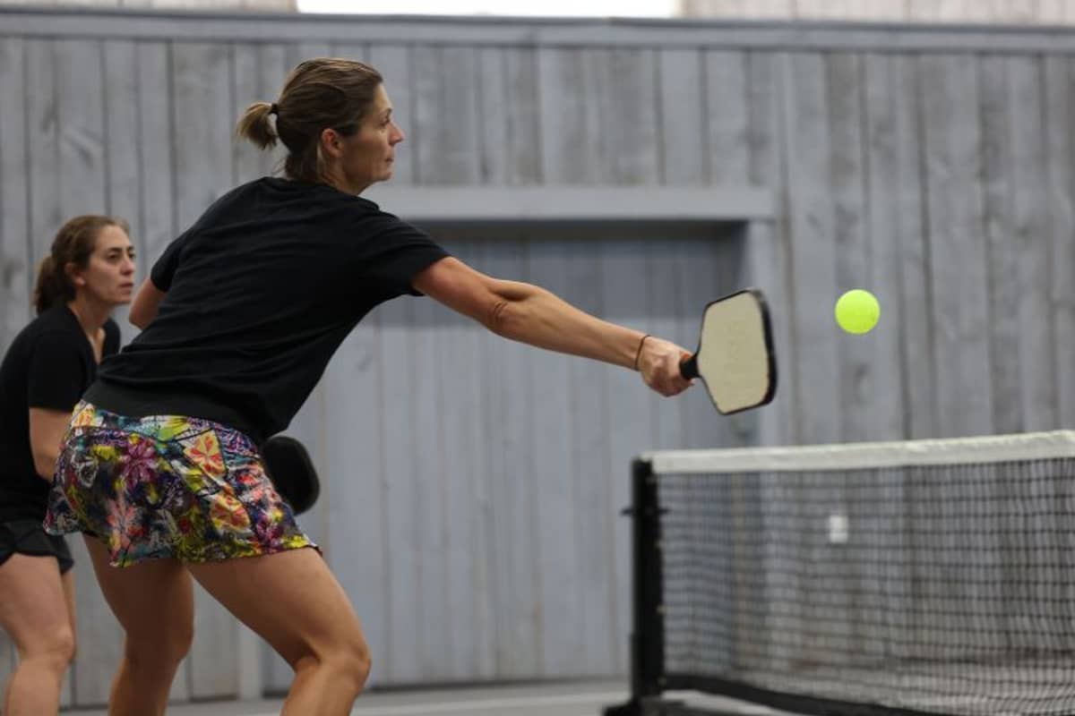 Two female pickleball players in an indoor court, with the foreground player making a forehand shot towards the net.