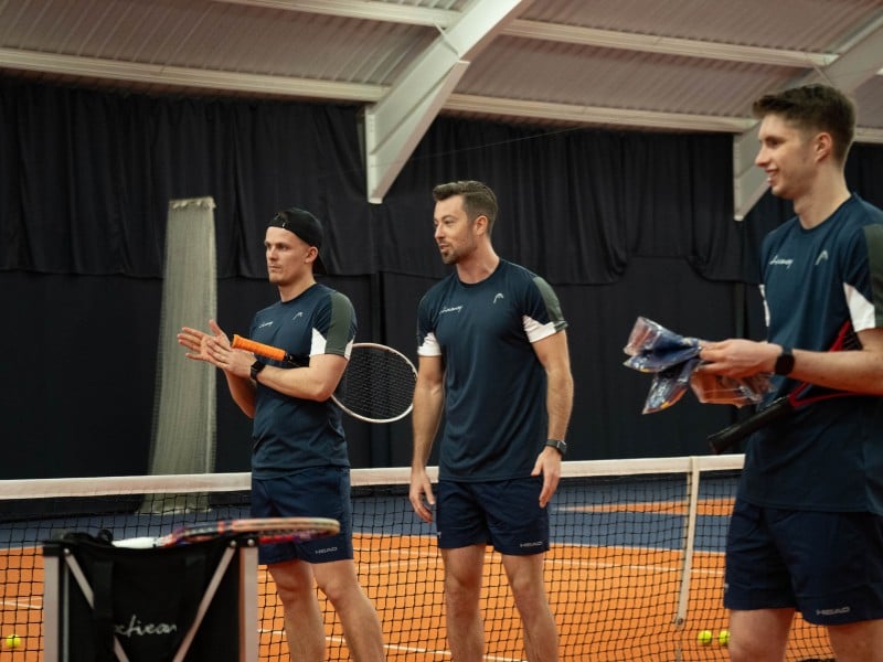 Three tennis coaches on an indoor court at David Lloyd Cheshire Oaks, wearing navy sports outfits and holding racquets.
