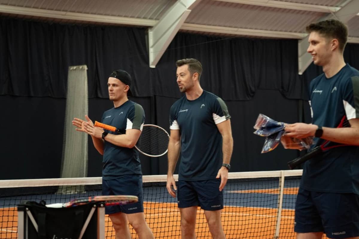 Three tennis coaches on an indoor court at David Lloyd Cheshire Oaks, wearing navy sports outfits and holding racquets.