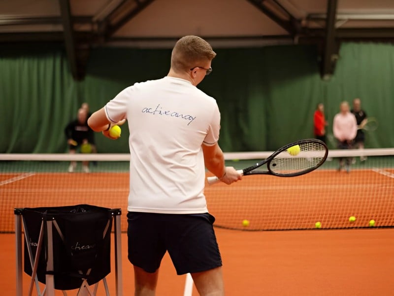 Tennis coach performing a forehand swing on an indoor court with students watching in the background at Cheshire Oaks.