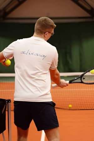 Tennis coach performing a forehand swing on an indoor court with students watching in the background at Cheshire Oaks.